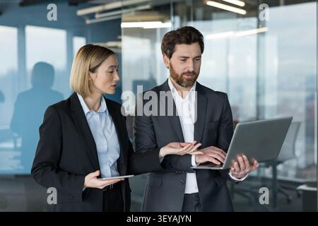 Deux collègues professionnels, un homme et une femme, debout ensemble dans un bureau contemporain, discutant de stratégie commerciale et utilisant un ordinateur portable et une tablette numérique pour le travail Banque D'Images
