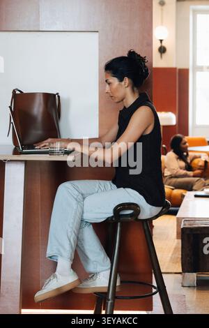 Vue latérale de la femme d'affaires concentrée travaillant sur son ordinateur portable tout en étant assis sur le tabouret à l'espace de coworking Banque D'Images