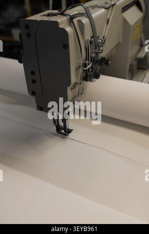 Vue rapprochée d'un mécanisme de machine à coudre industrielle cousant activement de grands rouleaux de tissu blanc, mettant en valeur la précision et l'efficacité dans le textile Banque D'Images