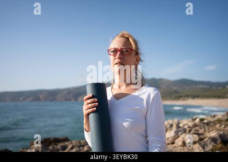 Vieille dame caucasienne portant des lunettes tenant un tapis de yoga sur la plage. Se préparer pour la séance de yoga matinale sur la rive de l'océan, promouvoir un mode de vie sain et l'idée de profiter de la retraite en vacances Banque D'Images