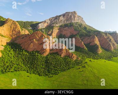 Prise de vue par drone à angle bas regardant de grandes falaises rouges multicolores recouvertes de buissons verts et d'un sommet de montagne gris vif Banque D'Images