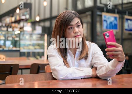 Portrait joyeux de style de vie d'une heureuse femme transgenre Isan asiatique du Sud-est dans ses années 40 doucement souriant tout en prenant selfie avec téléphone dans un restaurant t Banque D'Images