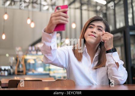 Portrait gai de style de vie d'une heureuse femme transgenre Isan d'Asie du Sud-est dans ses années 40 prenant selfie et faisant un geste de coeur avec la main dans un café. T Banque D'Images
