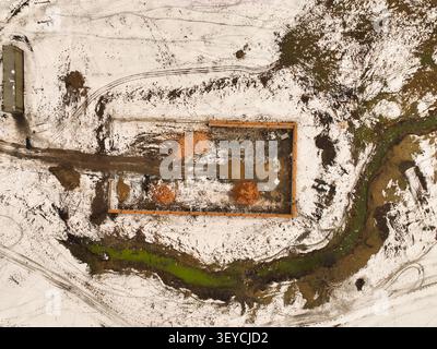 Vue aérienne directe d'un chantier de construction montrant une fondation en béton, des tas de bois d'œuvre et de terre, et des traces de boue dans la neige Banque D'Images