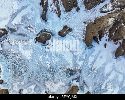 Vue aérienne détaillée de la surface gelée montrant des fissures bleues et des couches de glace blanche entourant des formations rocheuses sombres et dentelées Banque D'Images