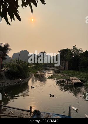 Lever de soleil doré brumeux sur un canal tropical bordé d'arbres luxuriants et de structures en bois sur pilotis. Aigrette blanche visible sur l'eau, Asie du Sud-est Banque D'Images