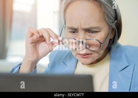 Femme mature stressée assise au travail, tenant des lunettes, ayant un problème de vision, des maux de tête ou un symptôme de migraine. Les yeux fatiguent la douleur concept Amal. Banque D'Images