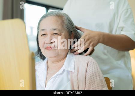 Joyeuse femme asiatique senior avec sa fille l'aide à faire ses cheveux, peignant ses cheveux en regardant un miroir à l'intérieur. Banque D'Images