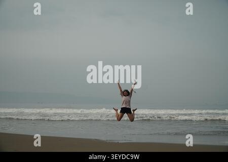Vue en pied d'une femme sautant les bras levés sur une plage de sable avec des vagues de l'océan sous un ciel nuageux en Indonésie Banque D'Images