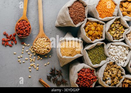 Grains assortis graines noix et fruits séchés dans des sacs en toile de jute avec cuillères en bois sur fond gris Flat Lay Banque D'Images