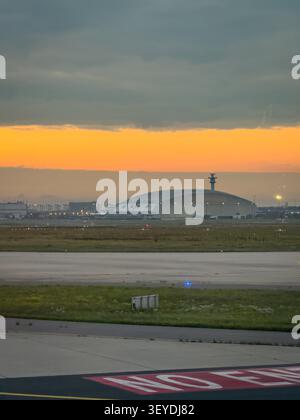 Francfort, Allemagne - 29 septembre 2025 : L'aéroport de Francfort au lever du soleil - tablier et voie de circulation avec des feux de bord bleus, un grand hangar d'entretien à l'horizon sous un ciel orange brillant Banque D'Images