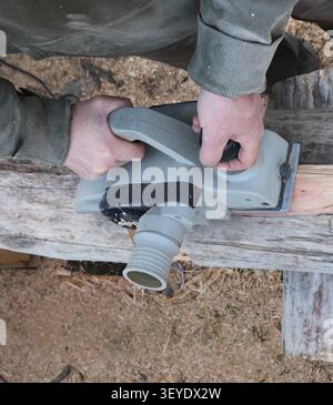 Paire de mains masculines guide avec assurance une défonceuse électrique grise le long d'une vieille bûche, enlevant la couche supérieure de bois ; vue en hauteur Banque D'Images