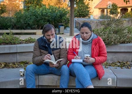 Un homme lisant un livre et une femme écrivant dans un cahier tout en étant assis côte à côte dans un parc d'automne. Banque D'Images