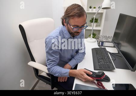Un homme d'affaires est assis à un bureau examinant un téléphone fixe tout en travaillant dans un espace de travail moderne. Banque D'Images
