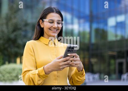 Jeune femme d’affaires portant des lunettes et une chemise jaune, souriant tout en interagissant avec un smartphone à l’extérieur, se connectant et communiquant dans un cadre d’affaires urbain contemporain Banque D'Images