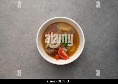 Soupe de boeuf copieux dans un bol blanc avec rondelles d'oignon, persil, tranches de tomate sur la surface de pierre grise Banque D'Images