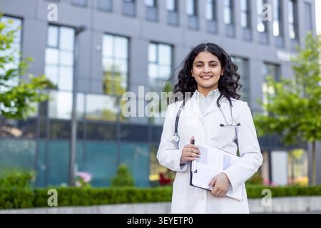 Jeune médecin indienne professionnelle souriant avec confiance à la caméra, portant une blouse de laboratoire blanche et un stéthoscope, tenant une planche à pince et un stylo tout en se tenant debout à l'extérieur devant un bâtiment moderne Banque D'Images