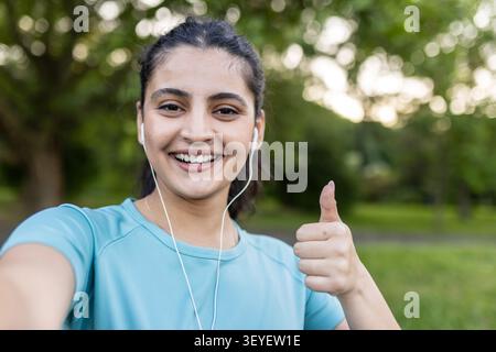 Jeune femme souriante tout en prenant un selfie à l'extérieur. Faire un geste du pouce levé. Prêt pour une séance d'entraînement et écouter de la musique avec des écouteurs. Symboliser la santé. Fitness. Réussite Banque D'Images