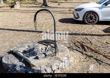Une photo en grand angle d'un feu de camp en pierre vide avec une structure de grille métallique, avec une voiture moderne blanche garée à proximité dans une forêt. Banque D'Images
