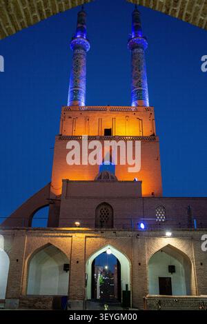 Un plan symétrique à angle bas encadré par une grande arche de briques, montrant le portail d'entrée lumineux chaud, surmonté de deux minarets en mosaïque bleue et violette Banque D'Images