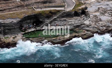 Vue aérienne de la piscine océanique de Bogey Hole creusée dans la roche avec des gens nageant dans l'eau verte claire, entourée de falaises accidentées et de vagues écrasantes, newca Banque D'Images