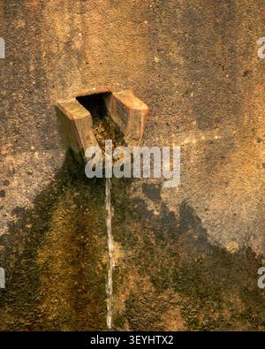 Gros plan d'un bec de fontaine en pierre altérée avec de l'eau claire qui coule sur un mur texturé dans une lumière naturelle chaude. Banque D'Images