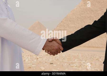 Gros plan poignée de main commerciale interculturelle entre arabe en thobe blanc et partenaire occidental approprié scellant un accord près des pyramides de Gizeh symbolisant Banque D'Images