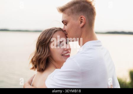 Un moment tendre entre un homme et une femme partageant un baiser sur le front près d'un plan d'eau calme pendant l'heure dorée Banque D'Images