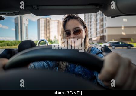 Jeune femme confiante conduisant une voiture décapotable à travers la ville, profitant de la balade. Style de vie urbain et conduite estivale heureuse. Banque D'Images
