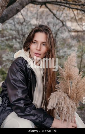 Femme veste jardin portrait d'une belle dame tenant de l'herbe de pampas dans un parc avec des arbres en fleurs au printemps Banque D'Images