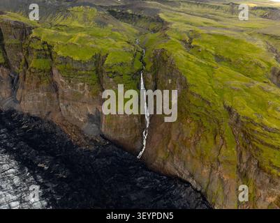Vue aérienne du glacier Solheimajokull avec une cascade descendant les falaises volcaniques abruptes couvertes de mousse à Myrdalshreppur, en Islande. Banque D'Images
