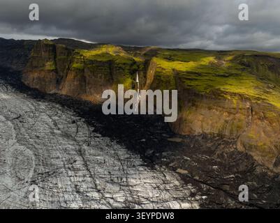 Vue aérienne du glacier Solheimajokull avec cendres volcaniques sombres, falaises moussues vertes vibrantes et une cascade sous un ciel nuageux spectaculaire i. Banque D'Images