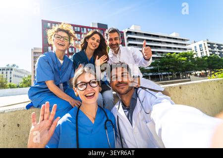 Équipe diversifiée de médecins et d'infirmières souriant et agitant pour un selfie à l'extérieur d'un bâtiment médical moderne, transmettant le travail d'équipe, l'unité et confiant, ami Banque D'Images