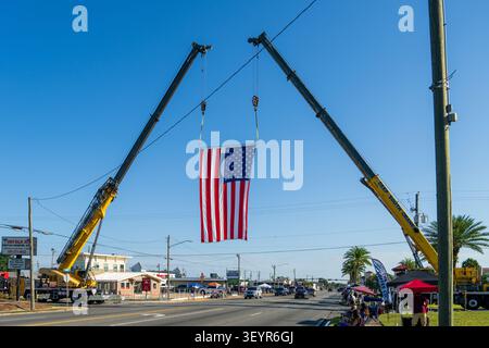 Lynn Haven, Floride, États-Unis. 4 juillet 2026. Deux grues Deep South suspendent un drapeau américain géant à travers Ohio Avenue sous un ciel bleu clair alors que les vendeurs installent des tentes ombragées et que les premiers spectateurs s'installent dans des chaises de pelouse le long de la route du défilé. Le drapeau ouvre Freedom by the Bay, la célébration annuelle du 4 juillet marquant le 250e anniversaire de l'Amérique, avec un défilé à 8h45 suivi d'une soirée au parc A.L. Kinsaul sur North Bay avec de la musique live de Taydee Marie et The Band Waleska, des food trucks et des feux d'artifice après le coucher du soleil. Crédit : Tosh Persons / Alamy Live News Banque D'Images