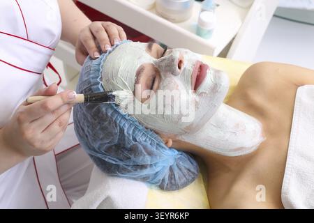 Cosmetician appliquant le masque facial à la face de belle jeune femme dans spa salon Banque D'Images