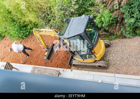 excavatrice travaillant activement et creusant sur un chantier de construction extérieur, entourée d'un tas de terre et d'un homme utilisant une pelle, capturée à partir d'une antenne Banque D'Images