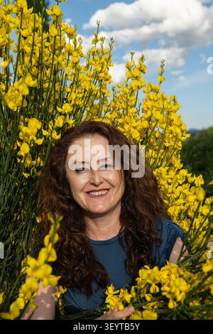 Un portrait d'une femme joyeuse aux cheveux bruns bouclés, souriant à la caméra devant des arbustes luxuriants en pleine floraison. Banque D'Images