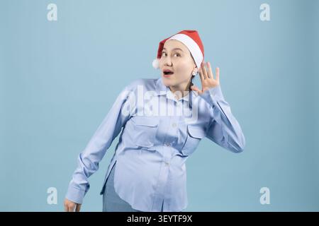 Femme enceinte touchant le visage portant chapeau de vacances en studio avec fond bleu Banque D'Images