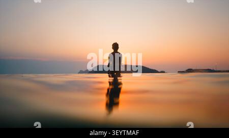 Enfant solitaire debout dans l'eau paisible regardant le coucher de soleil doré à l'horizon Banque D'Images