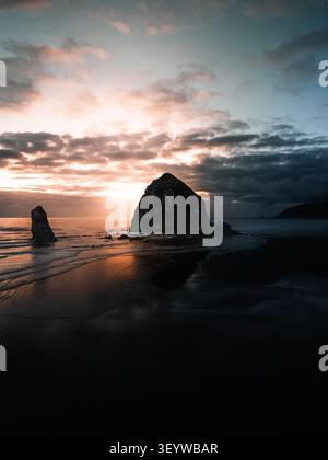 Vue aérienne de Haystack Rock au coucher du soleil avec une lumière dorée se reflétant sur le sable humide et des nuages spectaculaires au-dessus de l'océan Pacifique à Cannon Beach, Oregon Banque D'Images