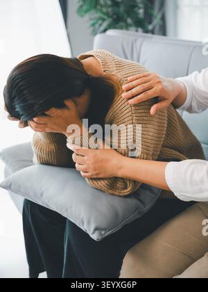 Triste jeune femme asiatique couvrant le visage et pleurant sur le canapé tandis qu'une autre femme la réconforte avec la main sur l'épaule, concept de dépression, de chagrin, de stress, un Banque D'Images