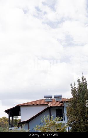 Une maison blanche de deux étages avec un toit de tuiles en terre cuite et des chauffe-eau solaires. Le bâtiment dispose d'un balcon et de grandes fenêtres entourées de verdure Banque D'Images