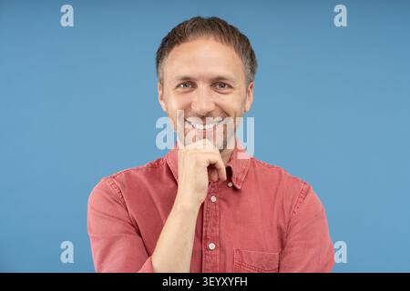 Portrait d'un homme d'âge moyen confiant souriant chaleureusement à la caméra avec la main reposant sur le menton sur fond de studio. Sourire attrayant, dents saines, positi Banque D'Images