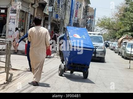 QUETTA, PAKISTAN - 05 juillet 2026 : dimanche, un ouvrier a été vu transporter un grand congélateur de marque Pepsi sur un chariot à main pour le réparer le long d'une route de cour très fréquentée à Quetta. La scène, capturée en plein jour, reflète l'agitation urbaine quotidienne, avec divers véhicules, dont un VUS argenté, naviguant dans la rue poussiéreuse. Des vitrines ornées d'affiches et d'un panneau « PRODEAL » bien en vue bordent la route, soulignant le dynamisme commercial de la région. Cette tâche routinière mais essentielle met en évidence les activités économiques en cours dans la région, cruciales pour les entreprises locales qui dépendent de la réfrigération fonctionnelle. Banque D'Images