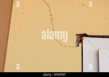 Vue rapprochée d'un mur extérieur jaune endommagé montrant une fissure proéminente et la texture rugueuse autour d'un cadre de fenêtre blanc avec isolati en mousse exposée Banque D'Images