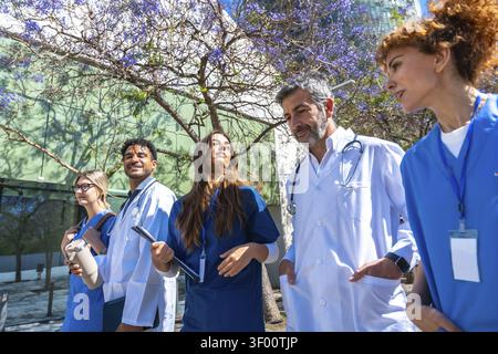 Groupe diversifié de professionnels de la santé et d'étudiants marchant à l'extérieur, discutant des concepts médicaux et du travail d'équipe pendant une journée ensoleillée à la clinique ou Banque D'Images