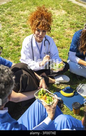Les membres de l'équipe médicale sont assis sur l'herbe, mangent des salades pour une pause déjeuner saine et relaxante, favorisant le bien-être et le renforcement de l'équipe en dehors du Banque D'Images