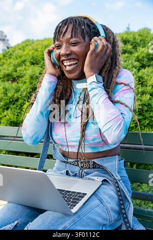 Joyeuse jeune femme portant des écouteurs rit tout en utilisant un ordinateur portable sur un banc de parc. Moment de style de vie en plein air avec tenue colorée, tresses et gr ensoleillé Banque D'Images