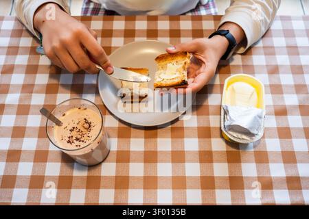 Vue aérienne d'une personne étalant du beurre sur du pain grillé sur une nappe à carreaux, avec une assiette, une boisson crémeuse et un bac à beurre, capturant une ambiance décontractée Banque D'Images