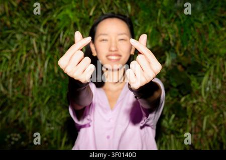 Souriante Gen Z femme asiatique debout dehors la nuit, atteignant la caméra et faisant des gestes de coeur de doigt avec les deux mains, avec un feuillage vert Banque D'Images